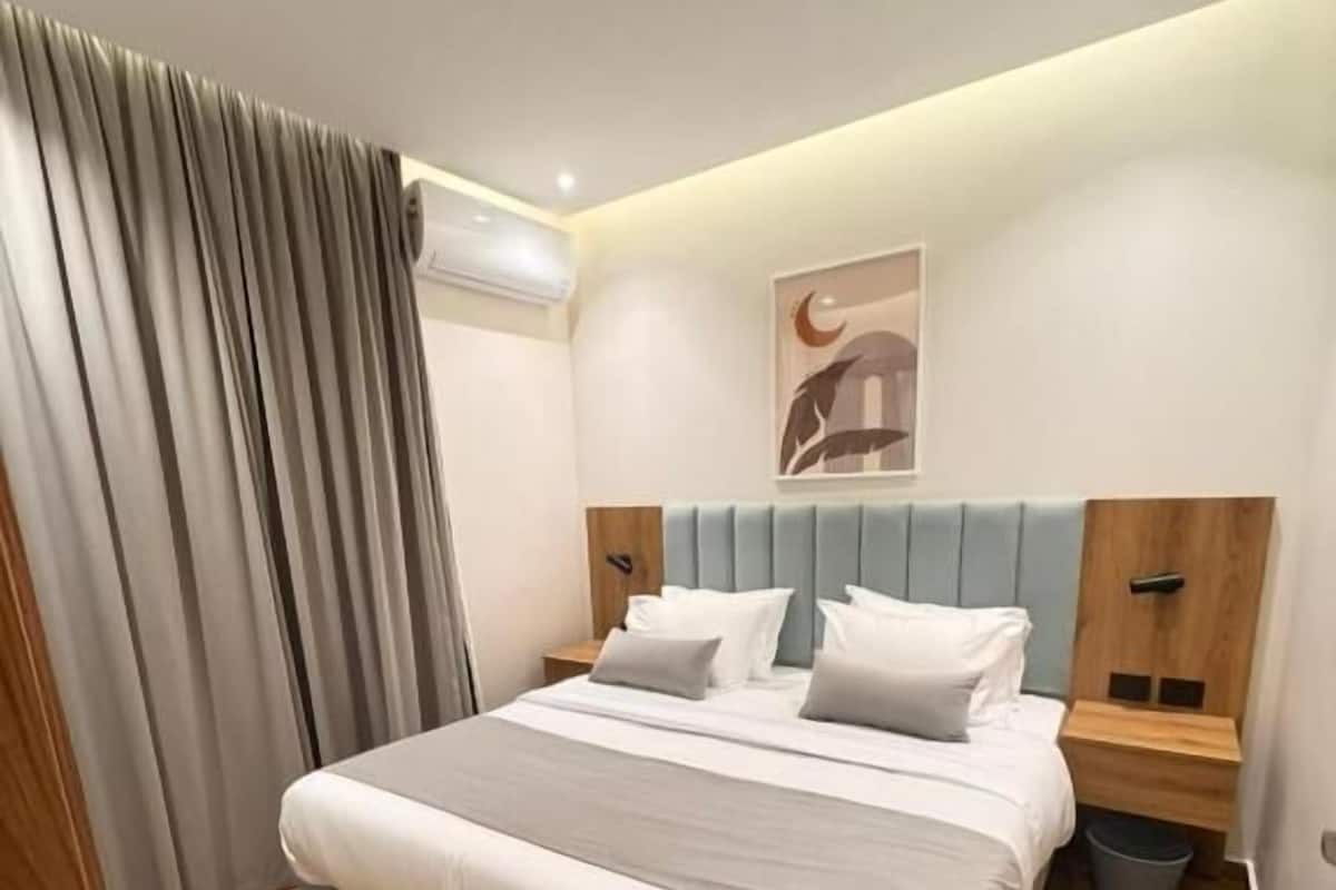 A neatly made bed with a headboard, a bedside table, a wall-mounted air conditioner, and a curtain.