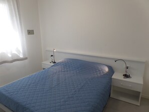 A bedroom with a single bed, a nightstand, a lamp, and a window with sheer curtains. - Modern Two-bed Flat With Shared Pool and hot tub (San Michele al Tagliamento)