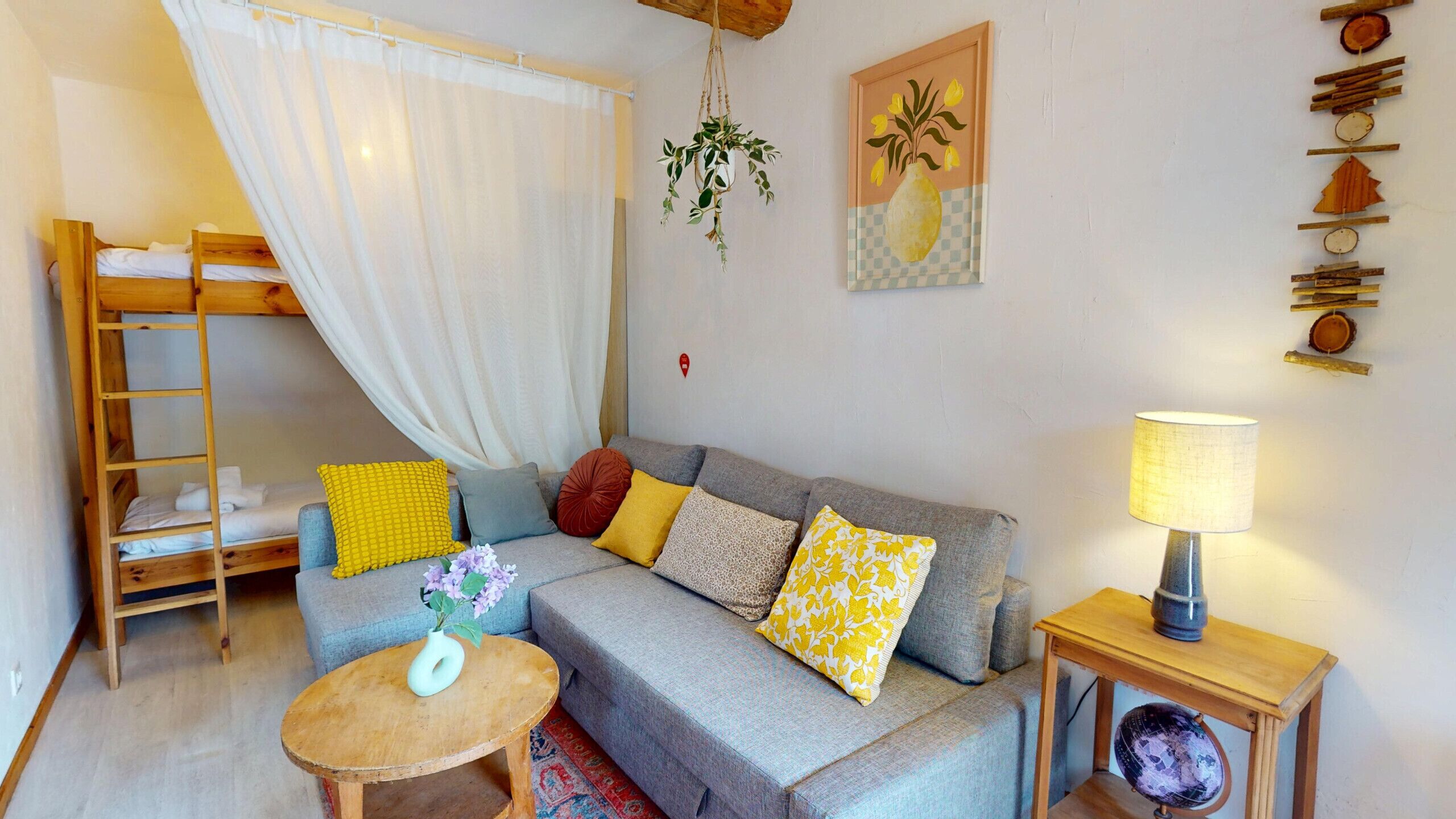 A cozy room with a bunk bed, a grey sofa with multiple cushions, a small round table with a vase, a lamp, and a wall-mounted plant.
