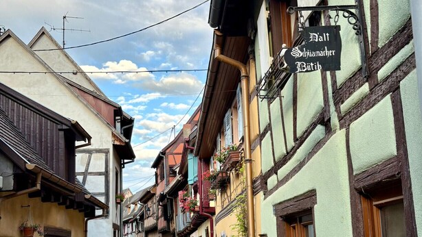 Eine Kopfsteinpflasterstraße gesäumt von traditionellen Fachwerkhäusern, ein Schild weist auf ein Restaurant hin, und der Himmel ist wolkenlos.