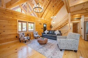 Ein rustikales Blockhaus-Interieur mit Holzboden, einem runden Couchtisch, einer Couch, gemusterten Sesseln, einer Treppe und Blick durch ein Fenster.