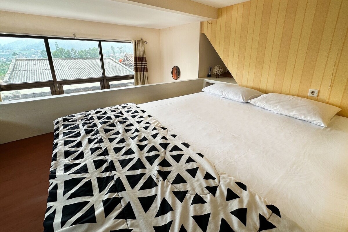 Une chambre d’hôtel avec un grand lit, une fenêtre donnant sur des immeubles, une tête de lit en bois et une table de chevet sur laquelle se trouve un vase.