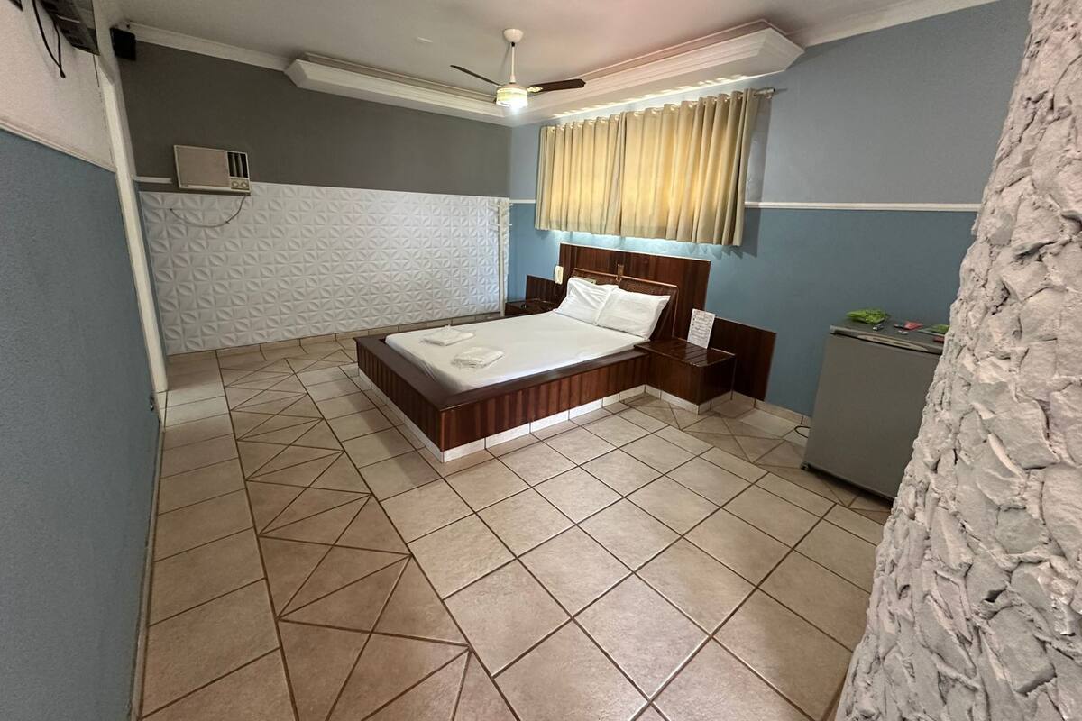 Un dormitorio con cama, mesitas de noche, ventilador de techo, ventana con cortinas y suelo de baldosas.
