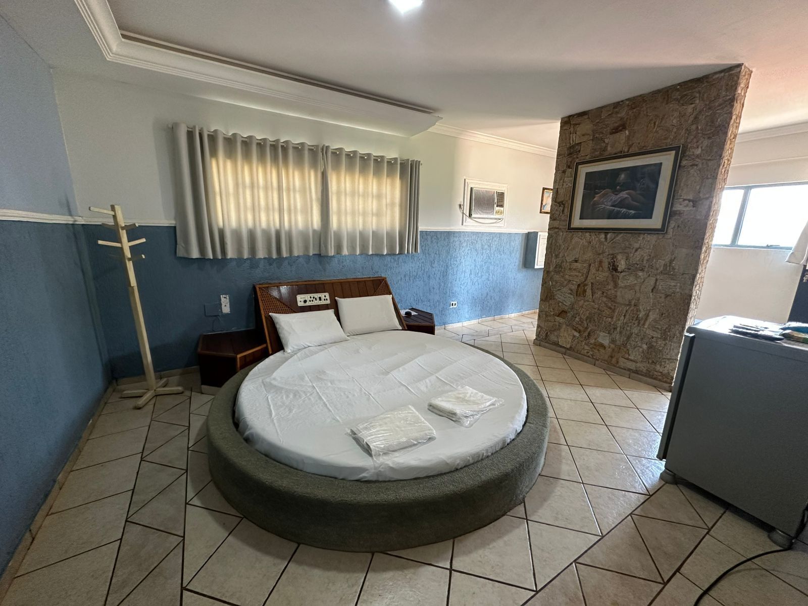 Una cama redonda con cabecera, una lámpara de pie, una ventana con cortinas, una pared de piedra decorativa y un cuadro enmarcado.
