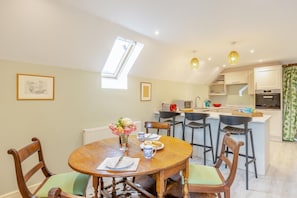 A dining area with a wooden table, chairs, a vase of flowers, and a kitchen in the background. - 1 Bed in Rimpton (oc-t38225) (Rimpton)