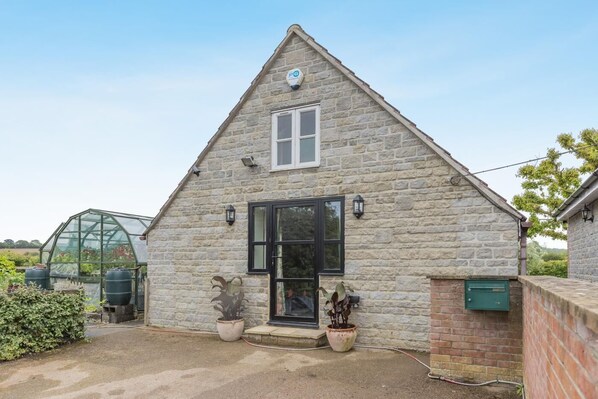 A stone building with a large window and a door, surrounded by potted plants and a greenhouse. - 1 Bed in Rimpton (oc-t38225) (Rimpton)