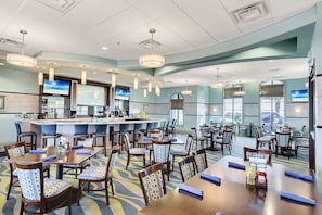 Une salle à manger d’hôtel avec de nombreuses tables et chaises, un bar et de grandes fenêtres.