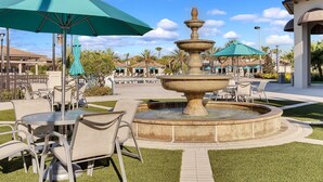 A fountain with multiple tiers, surrounded by a circular walkway. There are outdoor tables and chairs, and umbrellas providing shade. - Dream Vacation Villa Near Theme Parks with Pool! (Davenport)