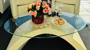 Una habitación con dos sillones blancos, una mesa de vidrio con un jarrón de flores, una botella de champán y un plato de pasteles.
