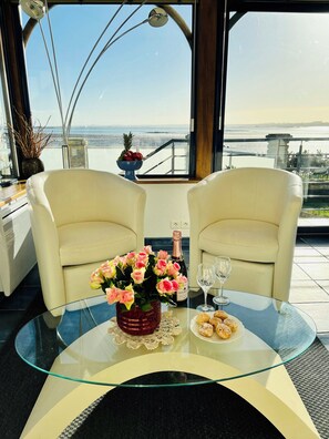 Ein Zimmer mit zwei weißen Sesseln, einem Glastisch mit Blumenvase, einer Champagnerflasche und einer Auswahl an Gebäck.