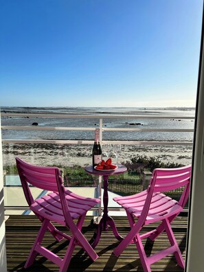 Ein Balkon mit zwei rosafarbenen Stühlen, einem kleinen Tisch mit Flasche und Gläsern und Blick auf den Strand.