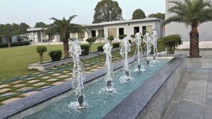 A water feature with multiple fountains in a rectangular basin, surrounded by a landscaped garden with palm trees and a building in the background. - THE FORESTAA RESORT (Haridwar)