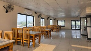 A spacious room with multiple wooden tables and chairs, large windows, ceiling fans, and an air conditioning unit. - THE FORESTAA RESORT (Haridwar)