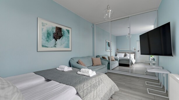 A modern hotel room with a bed, a sofa, a TV, and a large mirror. - Glow - A Cozy Studio in a Great Location (Sopot)