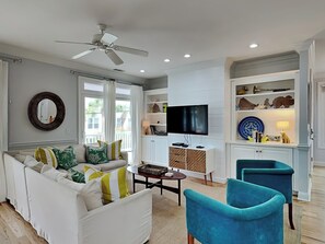 A living room with a sofa, a television, a ceiling fan, and a bookshelf. - Chu Cottage (Tybee Island)