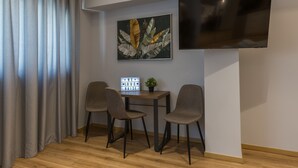 Une chambre moderne avec un plancher en bois, deux fauteuils gris, une petite table avec une plante et un panneau, un téléviseur à écran plat et une grande fenêtre avec des rideaux.