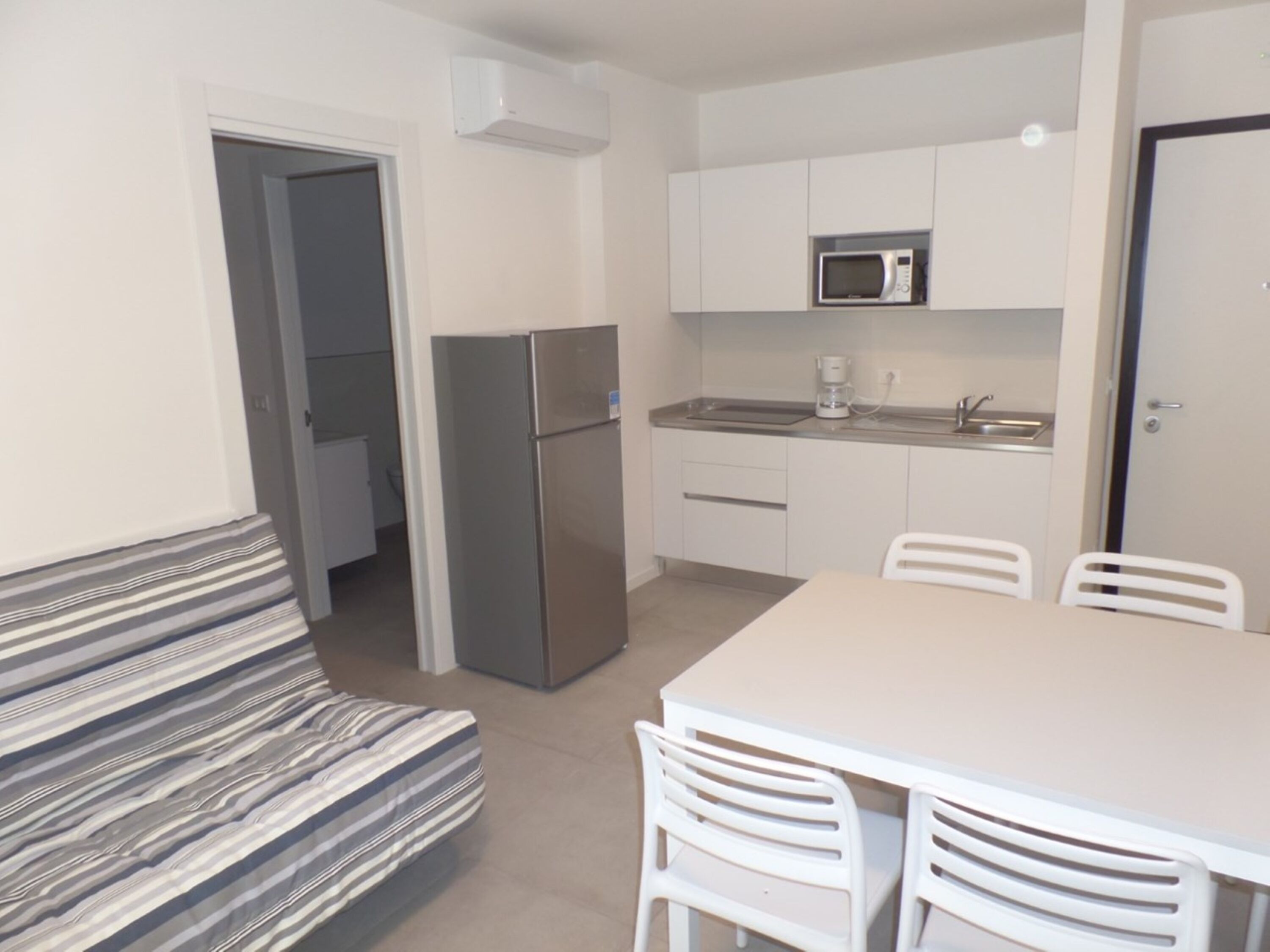 A compact kitchen with a refrigerator, microwave, and sink. There is a dining table with chairs and a striped bench.