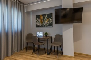 Ein modernes Zimmer mit Holzboden, zwei grauen Stühlen, einem kleinen Tisch mit Pflanze und Schild, einem Flachbildfernseher und einem großen Fenster mit Vorhängen.