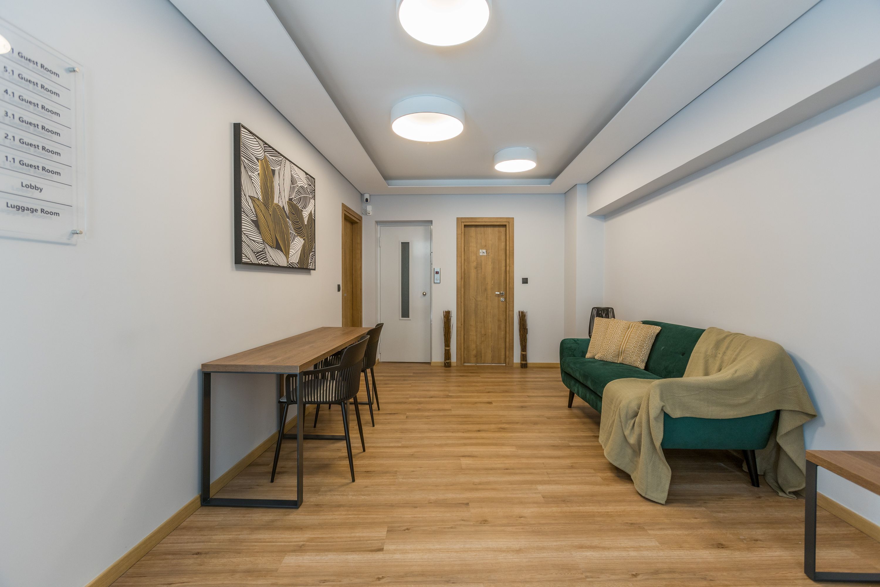Un couloir avec un plancher en bois, un canapé vert, une table en bois avec des chaises, et un panneau indiquant l’emplacement des chambres d’hôtes et du hall d’accueil.