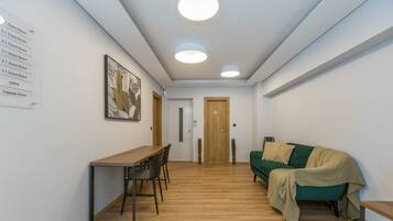 Un couloir avec un plancher en bois, un canapé vert, une table en bois avec des chaises, et un panneau indiquant l’emplacement des chambres d’hôtes et du hall d’accueil.