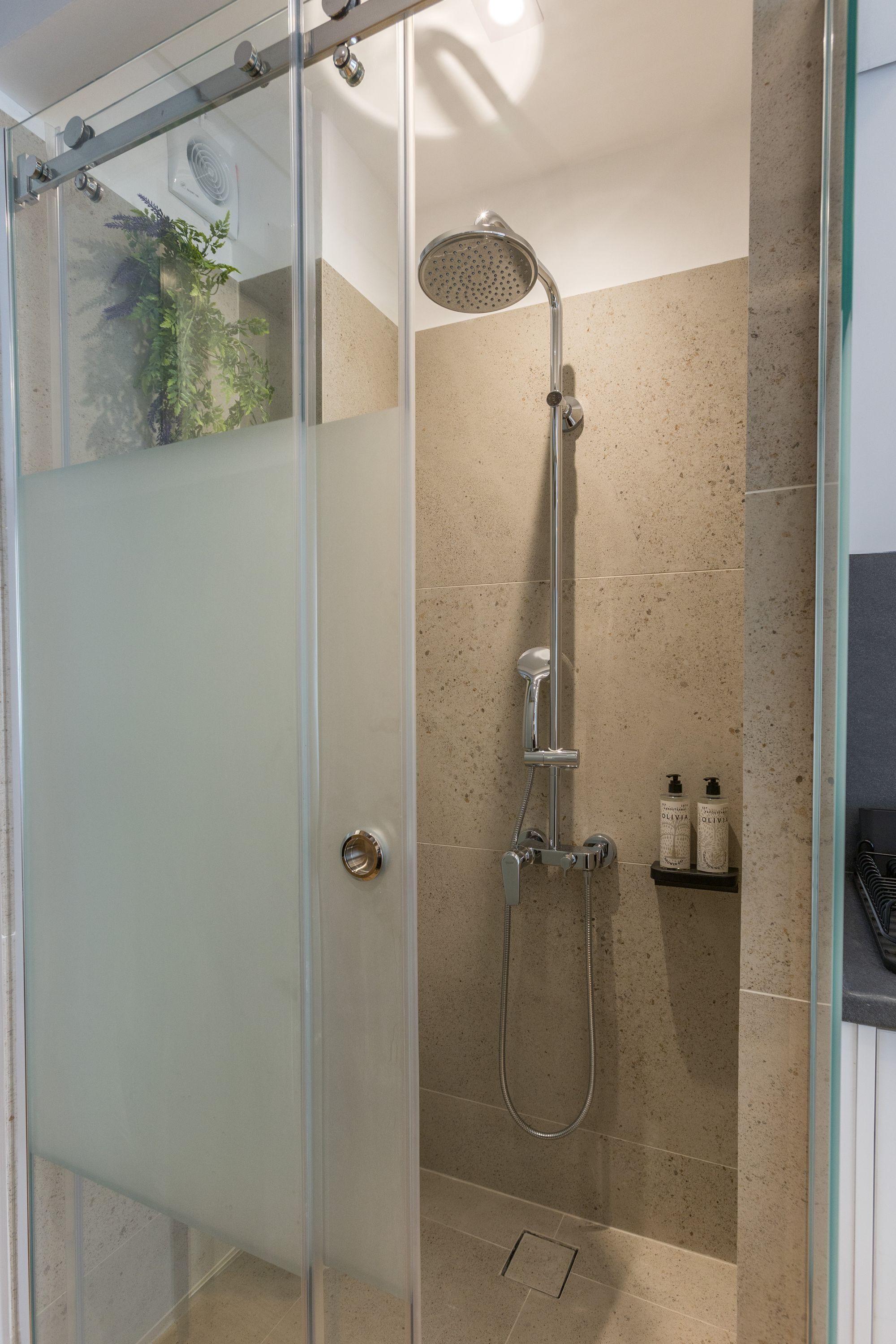 Une douche fermée par des parois en verre, dotée d’un pommeau de douche à main, d’un robinet mural et d’une petite étagère pour les articles de toilette.