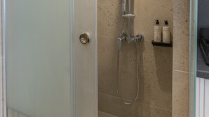 A glass-enclosed shower with a handheld showerhead, a wall-mounted faucet, and a small shelf with toiletries. - Junior Suite with balcony by Aethir Urban Stay (Athina)
