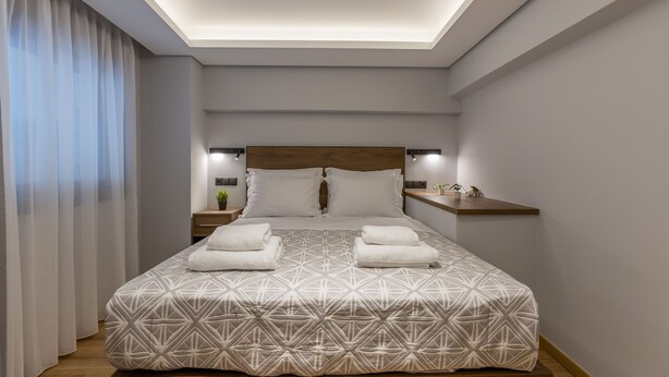 Ein modernes Hotelzimmer mit einem geometrisch gemusterten Bett, einem Holzkopfende, Nachttischlampen, einem Nachttisch mit Pflanze und einer eingebauten Ablage.