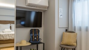 Ein modernes Hotelzimmer mit Holzboden, einem Flachbildfernseher, einer Deckenklimaanlage, einem kleinen Tisch mit Pflanze, einem Sessel mit Kissen und einem Bett mit gemustertem Überwurf.