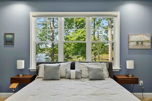 A bedroom with a large window, a bed with pillows, two bedside tables with lamps, and a wall with a sign and a picture.