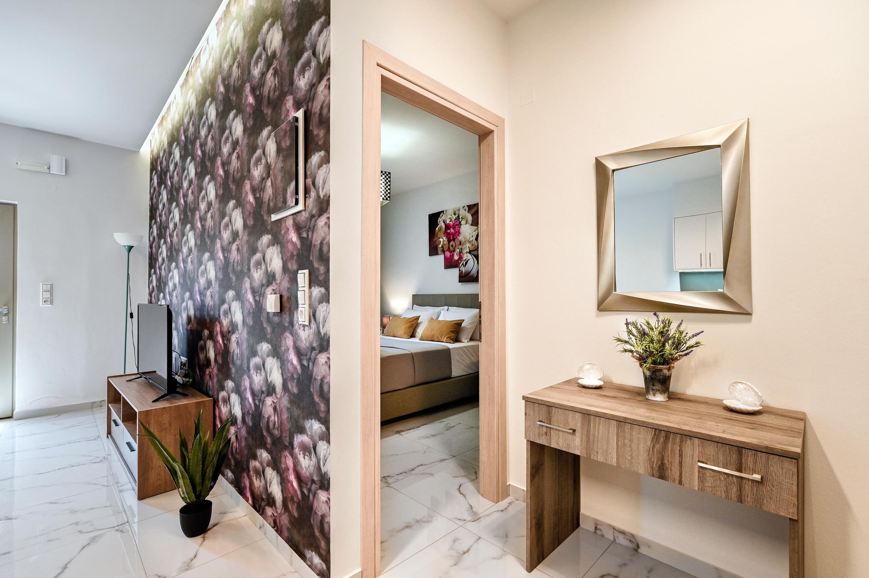 Ein modernes Hotelzimmer mit einem großen Spiegel, einer Holzkonsole, einer Topfpflanze und Blick in den Schlafbereich.