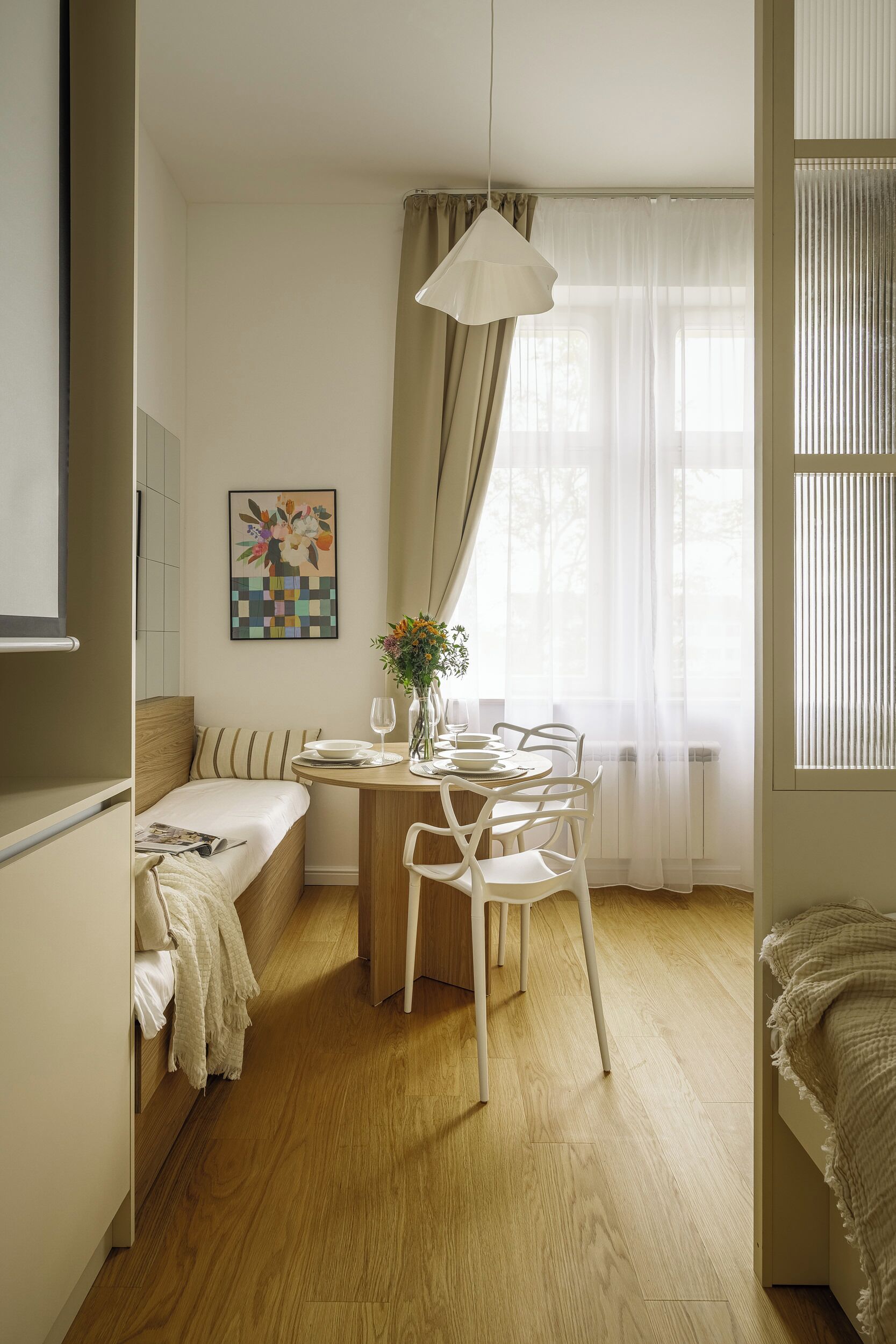 Et lille værelse med en seng, et spisebord med stole, en hængende lampe, et vindue med gardiner og et billede hængt på væggen.