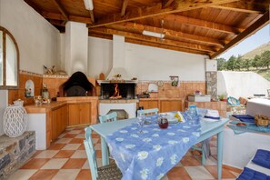 An outdoor kitchen with a built-in grill, a dining table with a blue floral tablecloth, and a stone fireplace.