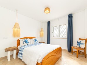 Ein Schlafzimmer mit einem hölzernen Bett, kariertem Bettbezug, zwei Nachttischen, einem Wandregal, einer Hängeleuchte, einem Fenster mit Vorhängen und einem Sessel.