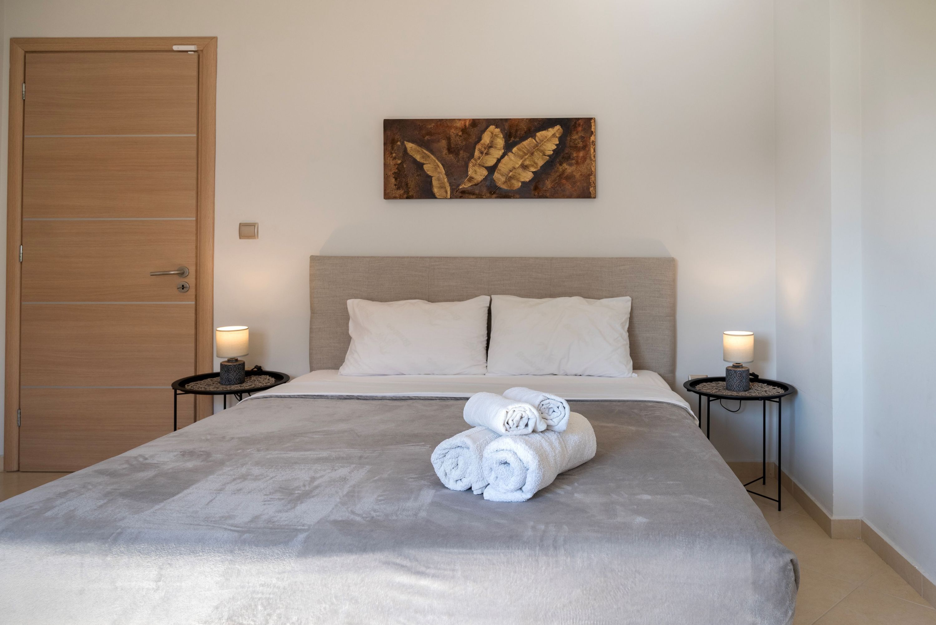 Ein ordentlich gemachtes Bett mit aufgerollten Handtüchern, einer Holztür und einem an der Wand hängenden Kunstwerk.