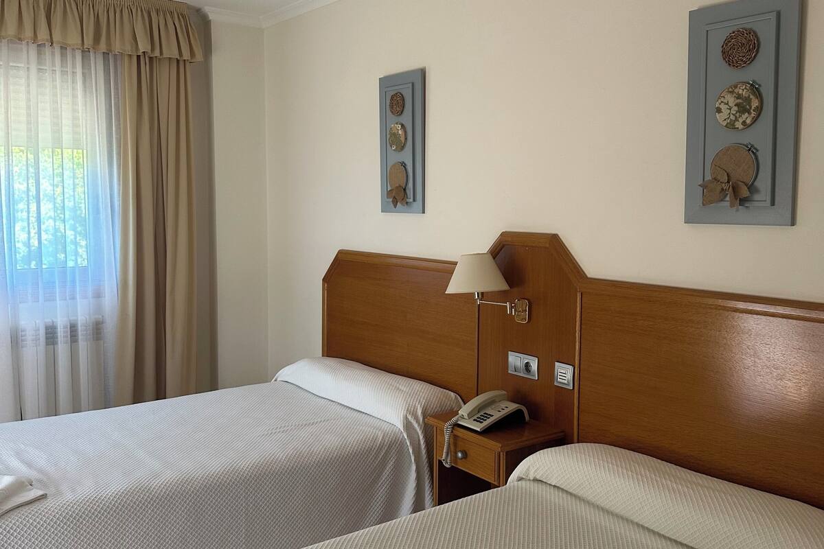 A hotel room with two beds, a bedside table with a lamp, a phone, and a wall-mounted artwork.
