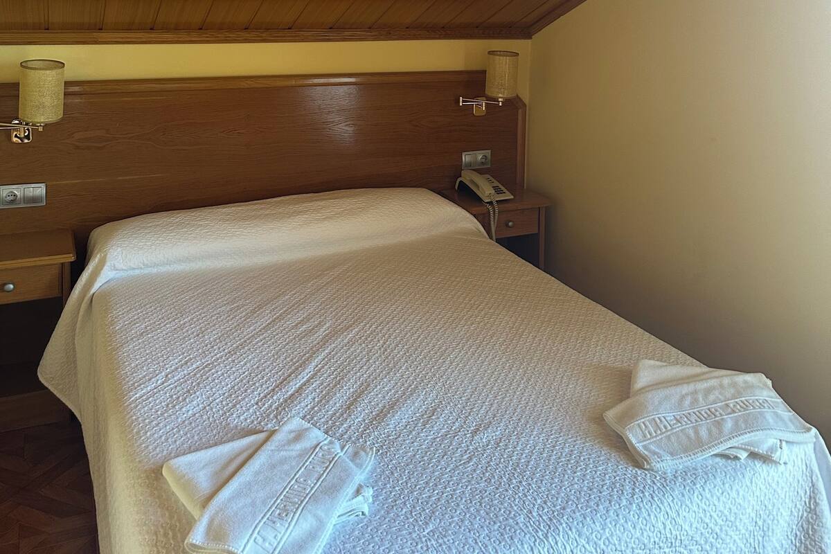 A hotel room with a wooden ceiling, a bed with a white textured bedspread, a nightstand with a lamp, and a wall-mounted telephone.