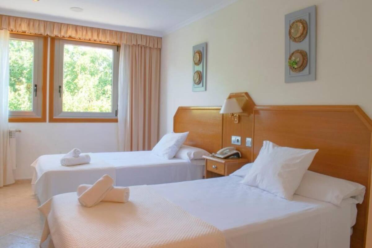 A hotel room with two beds, a wooden headboard, a bedside table with a lamp, and a window with curtains.