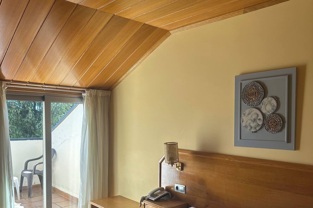 A bedroom with a bed, a bedside table with a lamp, a framed artwork, and a view of the outside.
