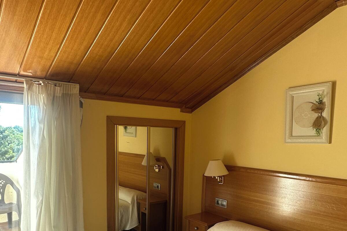 A hotel room with a wooden ceiling, a bed with a white bedspread, a nightstand with a lamp, and a door leading to a balcony.