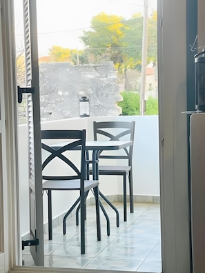 An open door leading to a balcony with a table and chairs, a lantern, and a view of greenery and a building in the background.