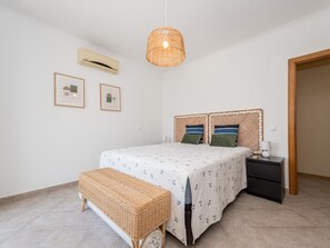 A bedroom with a bed, bedside table, bench, wall-mounted air conditioner, and two framed pictures. - CB LAG 10 2.E in Conceição de Tavira (Conceição de Tavira)