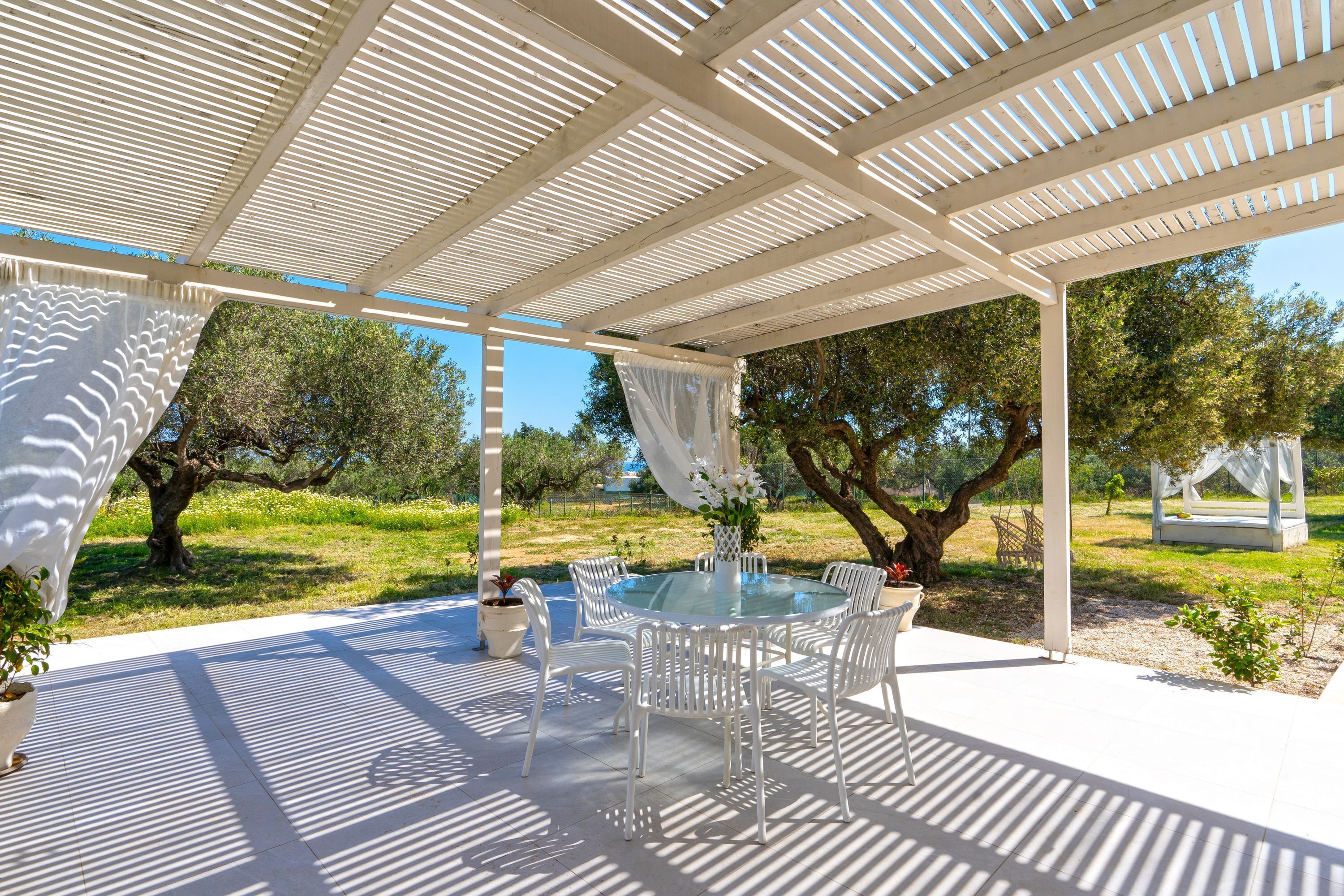 Uma área de refeições ao ar livre coberta, com mesa de vidro, cadeiras brancas e uma estrutura de pérgola.