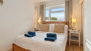 Een net opgemaakt bed met wit beddengoed en blauwe handdoeken, een houten hoofdbord, twee bedlampjes, een raam met gordijnen, en een klein wit nachtkastje met een mandje.