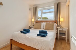Ein ordentlich bezogenes Bett mit weißer Bettwäsche und blauen Handtüchern, ein Holz-Kopfteil, zwei Nachttischlampen, ein Fenster mit Vorhängen und ein kleiner weißer Nachttisch mit einem Korb.