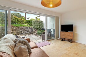 Ein Wohnzimmer mit einer Couch, einem Fernseher auf einem Holzboden und einer Glasschiebetür, die zu einem Außenbereich mit Steinmauer und Pflanzen führt.