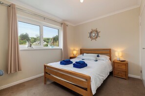 A bedroom with a wooden bed, two bedside tables with lamps, a window with curtains, and a wall decoration. - Smugglers Cove - Short Walk to Beach (Amroth)