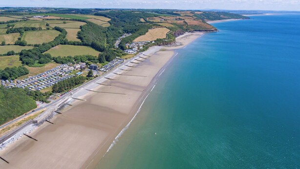Vista aérea de uma área costeira com uma extensa praia de areia, um estacionamento e uma fileira de edifícios à beira-mar.
