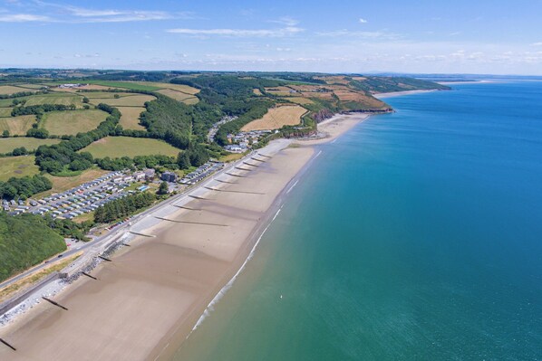 An aerial view of a coastal area with a long sandy beach, a parking lot, and a row of buildings along the shore. - Smugglers Cove - Short Walk to Beach (Amroth)