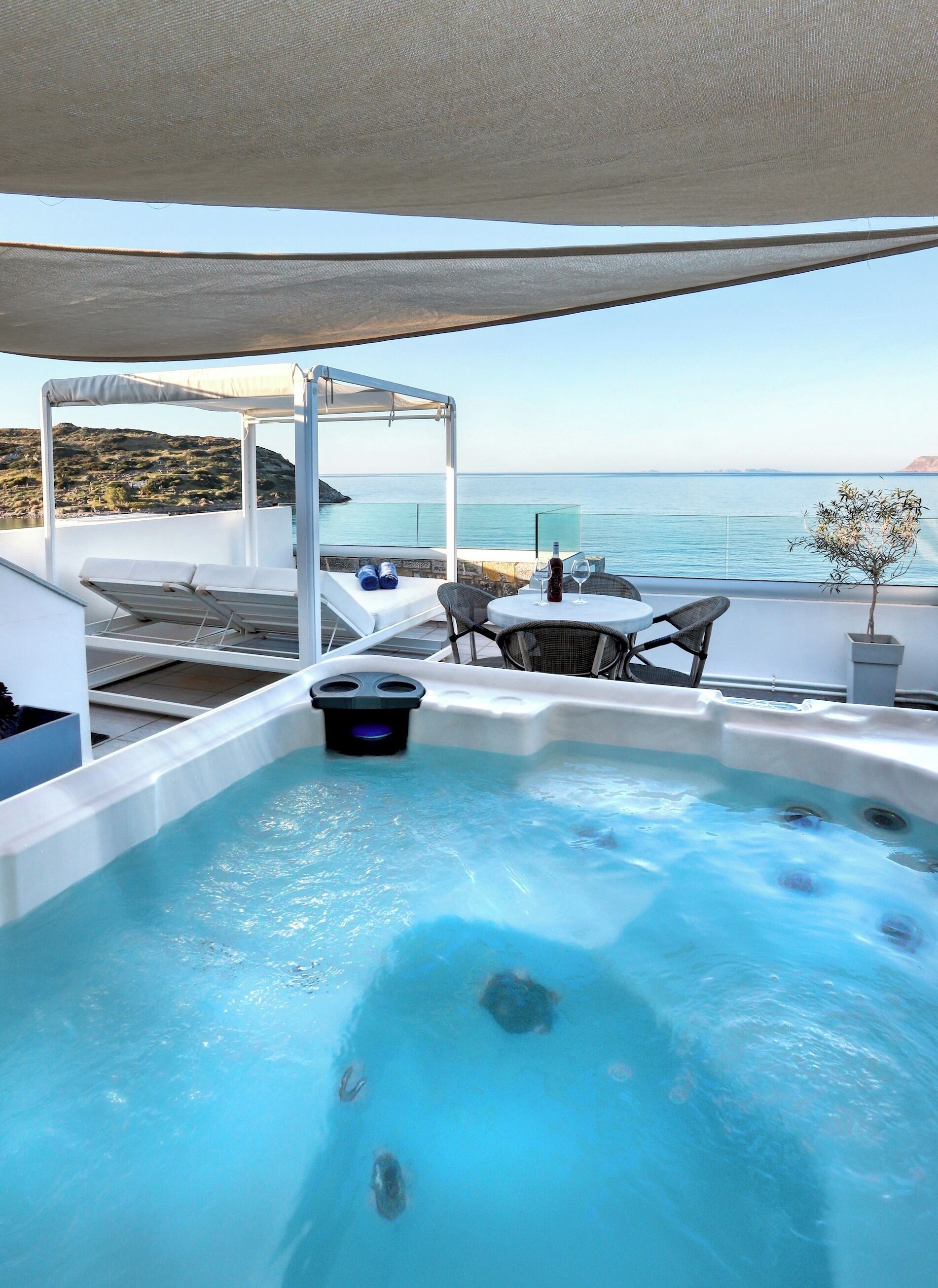 Un bain à remous avec vue sur la mer, un espace salon couvert et une petite table avec une bouteille et un verre.
