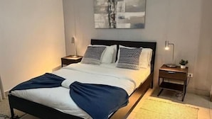 A modern bedroom with a bed, bedside tables, a wall-mounted artwork, and a chandelier. - Modern Ocean View 1BR in La Esperilla, Pool, Gym (Santo Domingo)